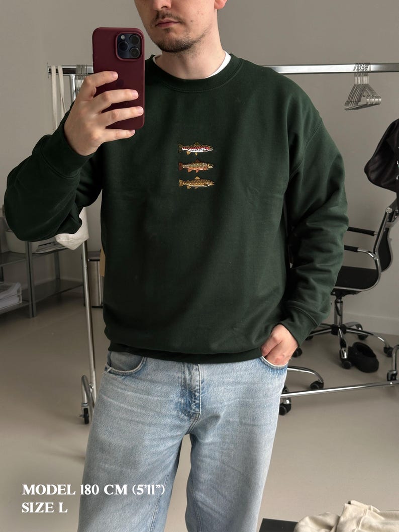 May include: A dark forest green crewneck sweatshirt featuring a small, centered embroidery of three stacked fish in shades of brown, orange, and white. The garment has long sleeves with ribbed cuffs and a ribbed neckline, paired with light blue denim jeans.