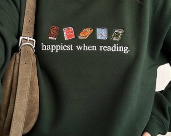 May include: A dark forest green crewneck sweatshirt featuring an embroidered row of five colorful books above the white text "happiest when reading." The sweatshirt is worn over a white collared shirt and paired with a brown suede shoulder bag.