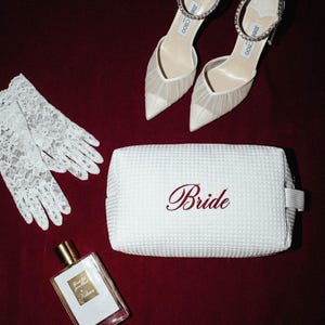 Puede incluir: Conjunto de accesorios de novia sobre fondo burdeos. Incluye guantes de encaje blanco, un neceser blanco con "Bride" bordado en rojo, un frasco de perfume y tacones color marfil con correas de tobillo de cristal.