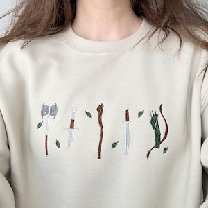 Puede incluir: Una sudadera de cuello redondo en color beige con un dise&ntilde;o bordado de armas de fantas&iacute;a que incluye un hacha de doble filo, una espada rota, un bast&oacute;n de madera, una daga y un arco con carcaj, todo rodeado de peque&ntilde;as hojas verdes bordadas.