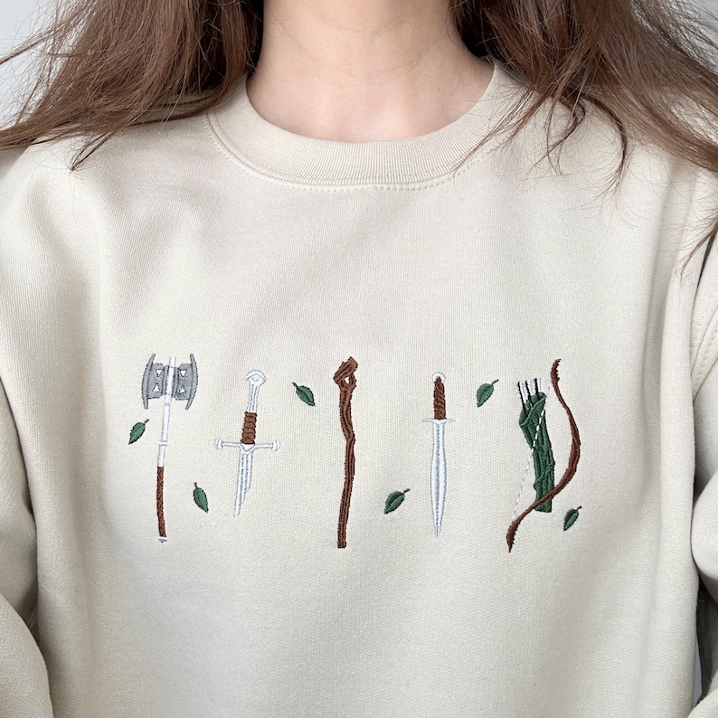 Puede incluir: Una sudadera de cuello redondo en color beige con un dise&ntilde;o bordado de armas de fantas&iacute;a que incluye un hacha de doble filo, una espada rota, un bast&oacute;n de madera, una daga y un arco con carcaj, todo rodeado de peque&ntilde;as hojas verdes bordadas.