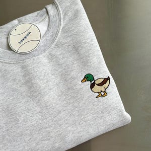 K&ouml;nnte beinhalten: Ein hellgraues Sweatshirt mit Rundhalsausschnitt und einer aufgestickten Stockente auf der Brust. Die Ente hat einen gr&uuml;nen Kopf, braune Fl&uuml;gel und gelbe F&uuml;&szlig;e. Ein rundes wei&szlig;es Etikett mit dem Text banagoya ist am Kragen befestigt.