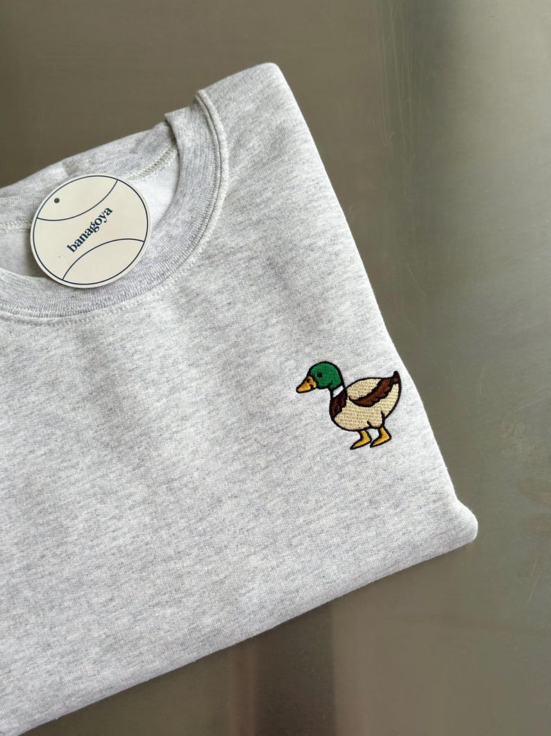 K&ouml;nnte beinhalten: Ein hellgraues Sweatshirt mit Rundhalsausschnitt und einer aufgestickten Stockente auf der Brust. Die Ente hat einen gr&uuml;nen Kopf, braune Fl&uuml;gel und gelbe F&uuml;&szlig;e. Ein rundes wei&szlig;es Etikett mit dem Text banagoya ist am Kragen befestigt.