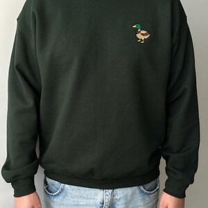 K&ouml;nnte beinhalten: Ein dunkelwaldgr&uuml;nes Sweatshirt mit Rundhalsausschnitt und einer kleinen, gestickten Stockente auf der linken Brust. Die Ente hat einen gr&uuml;nen Kopf, einen gelben Schnabel und einen braunen K&ouml;rper. Das Sweatshirt hat lange &Auml;rmel mit gerippten B&uuml;ndchen.