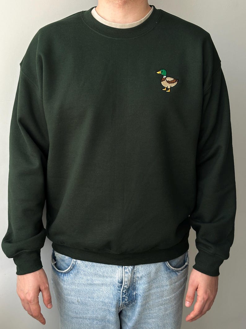 K&ouml;nnte beinhalten: Ein dunkelwaldgr&uuml;nes Sweatshirt mit Rundhalsausschnitt und einer kleinen, gestickten Stockente auf der linken Brust. Die Ente hat einen gr&uuml;nen Kopf, einen gelben Schnabel und einen braunen K&ouml;rper. Das Sweatshirt hat lange &Auml;rmel mit gerippten B&uuml;ndchen.
