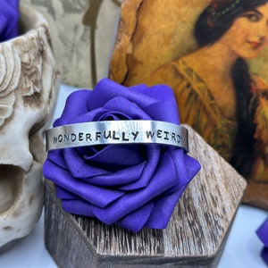 May include: A silver metal cuff bracelet with the words "WONDERFULLY WEIRD" stamped across the front. The bracelet is displayed on a purple satin rose, with a skull and vintage artwork in the background.