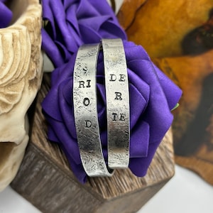May include: Two silver bracelets with the words "Ride" and "Ride" engraved on them. The bracelets are on a purple background.