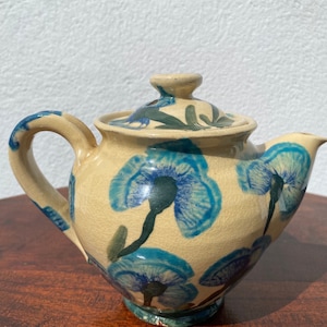 May include: A cream-coloured ceramic teapot with a blue and green floral design. The teapot has a lid and a spout.