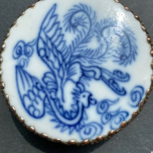 May include: A circular ceramic piece featuring a blue phoenix design on a white background. The edge is trimmed with a copper-coloured metal. The phoenix is shown with detailed wings and cloud motifs. The piece is likely decorative.
