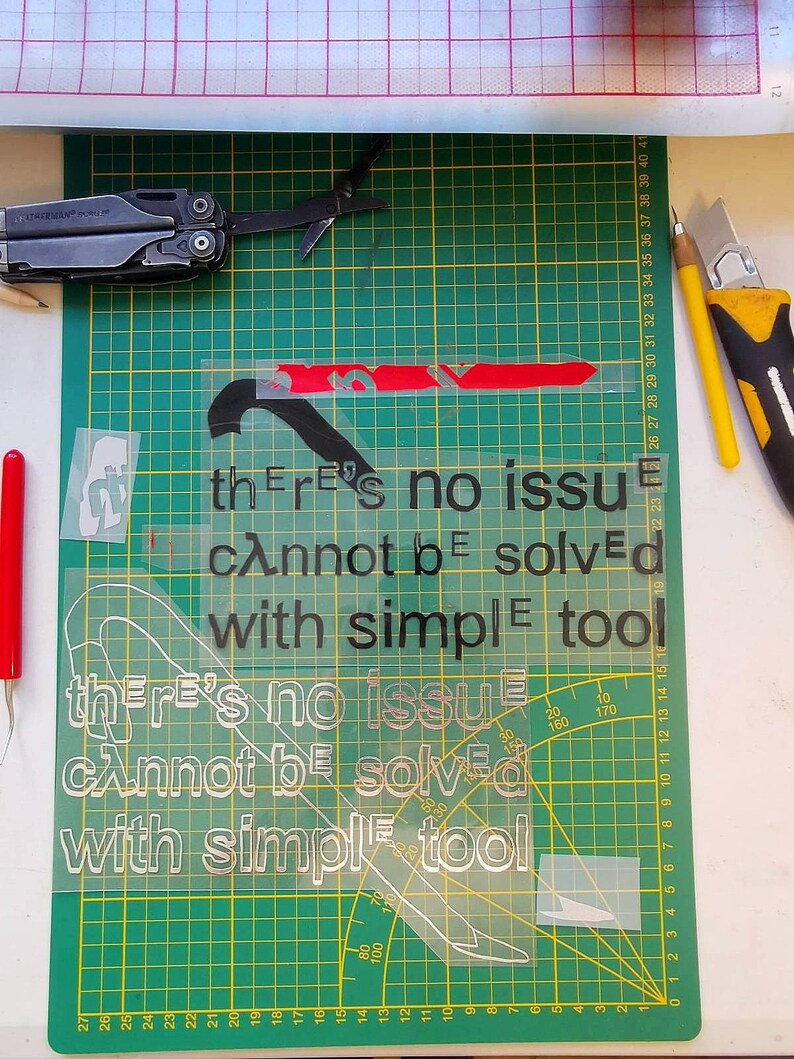 May include: A green cutting mat with a white stencil on top. The stencil has the text "there's no issue that cannot be solved with simple tools" repeated twice. The text is in black and the background of the stencil is white.