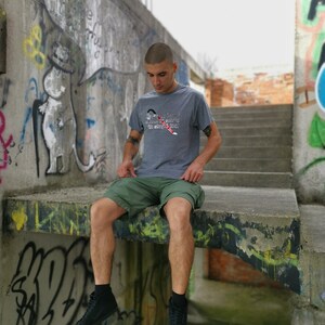 May include: A young person wearing a grey t-shirt with the text "My passion is showing up with empty pockets" in white and red. They are wearing green shorts and black high-top sneakers. The person is sitting on a concrete ledge with graffiti in the background.