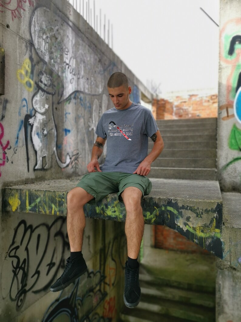 May include: A young person wearing a grey t-shirt with the text "My passion is showing up with empty pockets" in white and red. They are wearing green shorts and black high-top sneakers. The person is sitting on a concrete ledge with graffiti in the background.