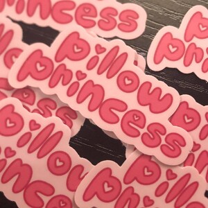 May include: A collection of pink stickers with the words "Pillow Princess" in a bubble font. Each sticker has a white border and small heart details. The stickers are scattered on a dark surface.