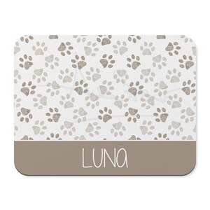 May include: A white rectangular pet placemat with a brown paw print pattern and the name "LUNA" printed in a brown rectangle at the bottom.