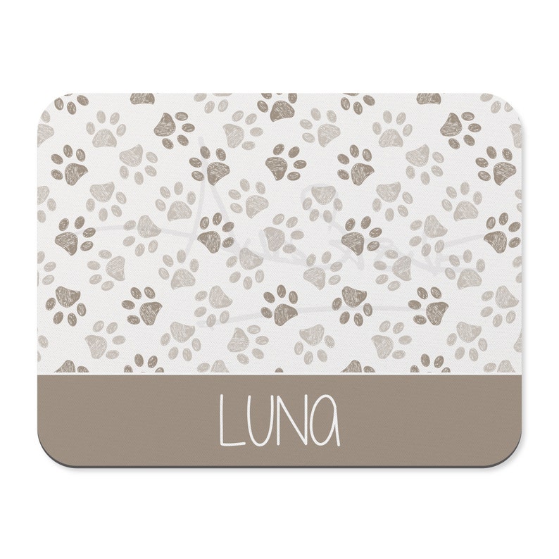 May include: A white rectangular pet placemat with a brown paw print pattern and the name "LUNA" printed in a brown rectangle at the bottom.