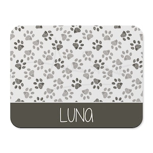 May include: A white rectangular pet mat with a gray paw print pattern and a dark gray border. The name "LUNA" is printed in white on the border.