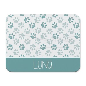 May include: A teal blue rectangular pet placemat with a white background and a pattern of teal blue paw prints. The name "LUNA" is written in white script at the bottom of the placemat.
