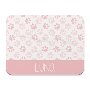 May include: Pink mouse pad with a white background and a pattern of pink paw prints. The bottom of the mouse pad has a solid pink band with the name "LUNA" written in white.