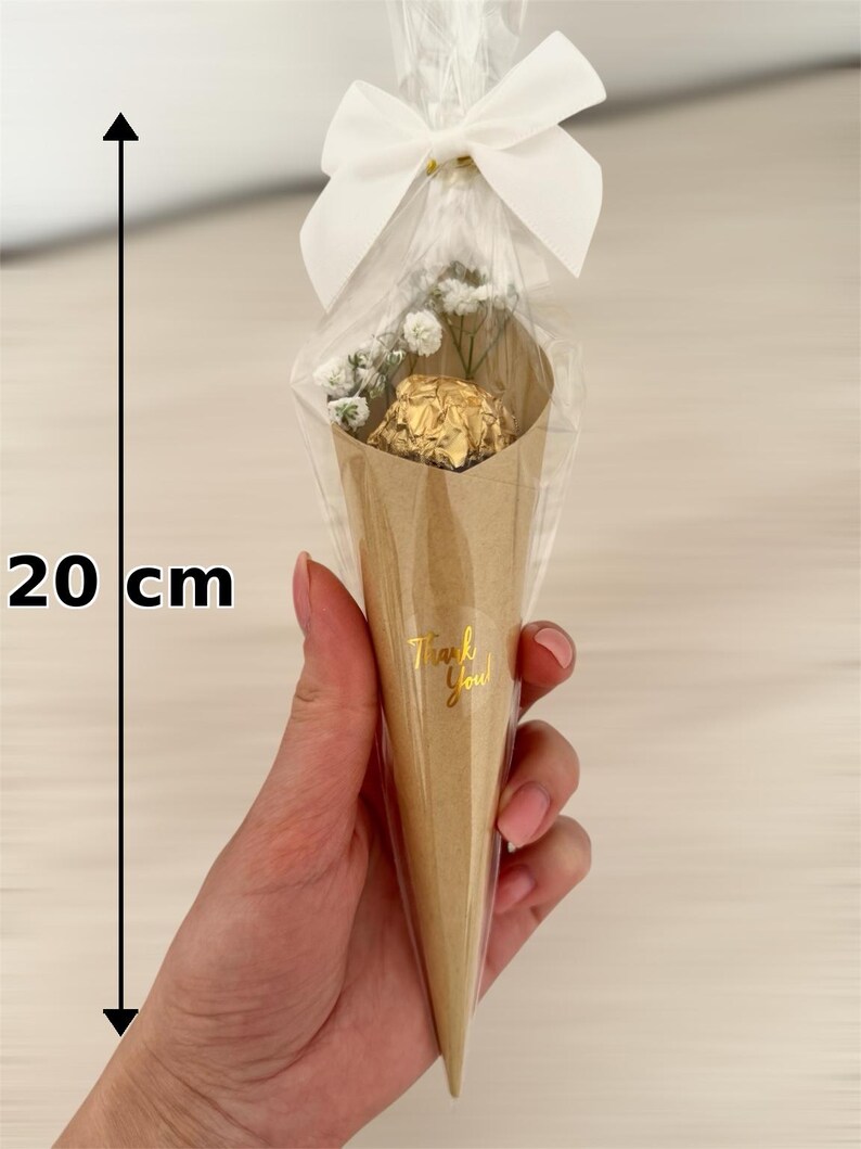 May include: A cone-shaped gift package, approximately 8 inches tall, made of brown paper, containing a gold-wrapped chocolate and small white flowers. The package is tied with a white bow and cellophane. The text "Thank You!" is printed on the cone.