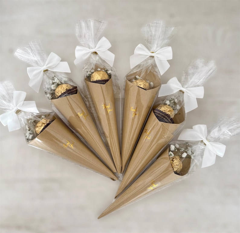 May include: Six cone-shaped party favors. Each favor is made of brown paper, filled with a chocolate candy and baby's breath, and wrapped in clear cellophane. White bows secure the cellophane. The words "Thank You" are printed in gold.