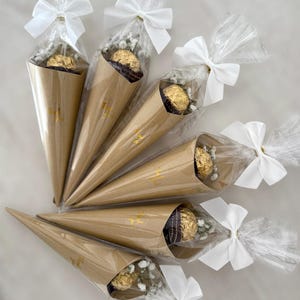 May include: Six brown paper cone-shaped packages tied with white bows. Each cone contains a gold-wrapped chocolate and small white flowers, wrapped in clear cellophane. Gold text is visible on the cones.