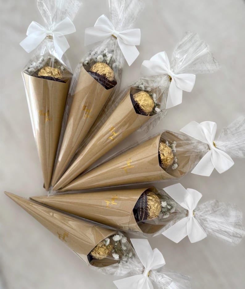 May include: Six brown paper cone-shaped packages tied with white bows. Each cone contains a gold-wrapped chocolate and small white flowers, wrapped in clear cellophane. Gold text is visible on the cones.