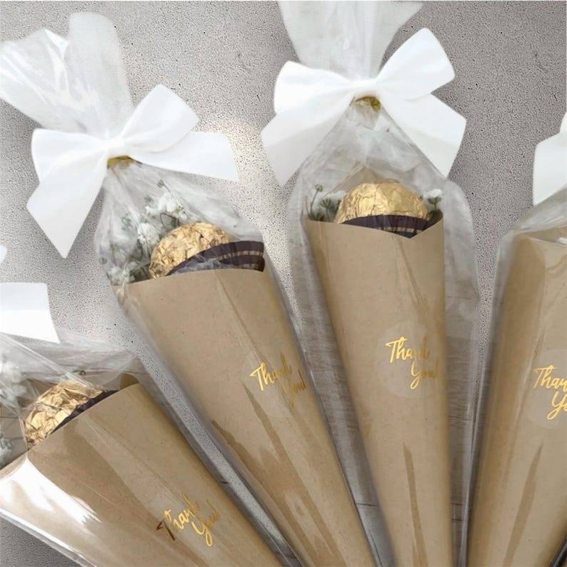 May include: Several cone-shaped gifts wrapped in brown paper and clear cellophane. Each gift contains a gold-wrapped chocolate and small white flowers. A white bow secures the cellophane. The words "Thank You" are printed on the paper.