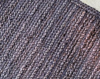 May include: Close-up of a dark gray woven jute rug. The rug has a textured surface with visible fibers and a slightly uneven color distribution, with hints of brown and tan. The rug's corner is visible.