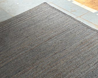 May include: A rectangular, grey jute rug with a woven texture. The rug's edges are neatly finished with a dark border. The rug is placed on a light-coloured stone surface, with a portion of a brown surface visible.
