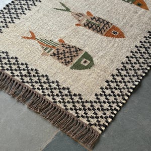 May include: A woven rug featuring a beige base with a repeating pattern of stylized fish in shades of orange, green, and brown. The rug is bordered by a black and white geometric pattern and finished with long, brown fringe along the short edge.
