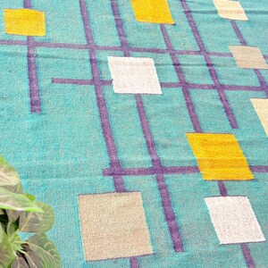 Könnte beinhalten: Türkisfarbener Teppich mit einem Gittermuster aus lila Linien und Quadraten in Gelb, Weiß und Beige. Der Teppich hat eine strukturierte Optik und ein geometrisches Design, passend für die Inneneinrichtung.