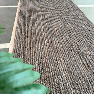 Puede incluir: Una alfombra de fibra natural hecha de yute, un material sostenible y duradero. La alfombra tiene una textura tejida y un color marrón claro.