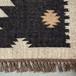 May include: Close-up of a woven rug with a geometric pattern. The rug features a black and off-white design with a tan accent. The bottom edge has a fringe. This rug is suitable for home decor.