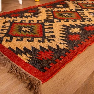 Könnte beinhalten: Ein roter, schwarzer und beiger Teppich mit Fransen. Der Teppich hat ein geometrisches Muster mit einem Rautenmuster.
