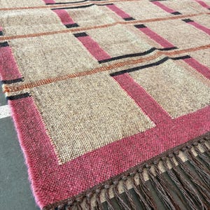 Könnte beinhalten: Ein gewebter Teppich mit einem geometrischen Muster in Braun-, Rosa- und Schwarztönen. Der Teppich hat einen Fransenrand.