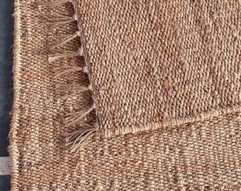 May include: A close-up of a natural fiber rug with a woven texture. The rug is made of a light brown material and has a fringe border.