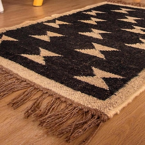 Puede incluir: Una alfombra tejida en negro y beige con un patrón geométrico y flecos en los bordes. La alfombra tiene un diseño moderno y minimalista.