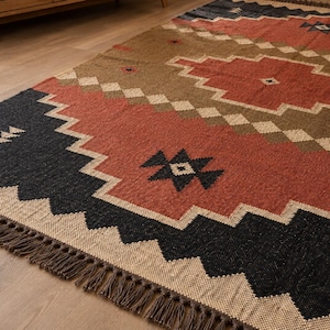 Könnte beinhalten: Ein rechteckiger Teppich mit einem geometrischen Muster aus großen Rauten und Zickzack-Bordüren. Die Farbpalette umfasst Terrakotta, Anthrazit und Beige. Der Teppich hat eine gewebte Struktur und ist an den Kanten mit kurzen Fransen versehen.
