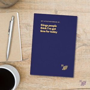 May include: A navy blue notebook with gold text reading "MY LITTLE NOTEBOOK OF... things people think I've got time for today." A silver pen and a coffee cup are on a wooden surface.