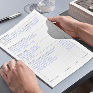 May include: A project plan document with blue text is held by two hands. A silver and black pen rests on the grey surface next to the document. The document includes sections for objectives, resources, and action steps.