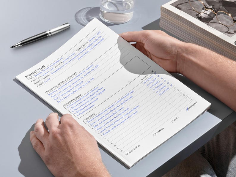May include: A project plan document with blue text is held by two hands. A silver and black pen rests on the grey surface next to the document. The document includes sections for objectives, resources, and action steps.