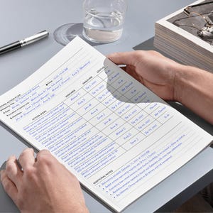 May include: A meeting action plan document with handwritten notes in blue ink. The document is held by a person and includes sections for action steps, deadlines, and assignees. A pen and a glass of water are on the table.