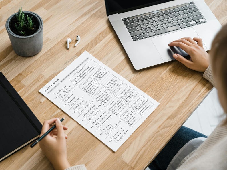 May include: A wooden desk with a laptop, a weekly work plan, a potted succulent, a notebook, and wireless earbuds. A person is using the laptop and writing on the work plan with a pen. The work plan is filled with handwritten notes.