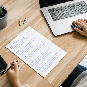 May include: A workspace scene featuring a wooden desk with a laptop, notebook, and a document with text. A small potted plant and wireless earbuds are also on the desk. A person's hand is using the laptop's touchpad and another hand is holding a pen.