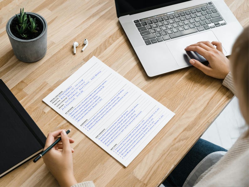 May include: A workspace scene featuring a wooden desk with a laptop, notebook, and a document with text. A small potted plant and wireless earbuds are also on the desk. A person's hand is using the laptop's touchpad and another hand is holding a pen.
