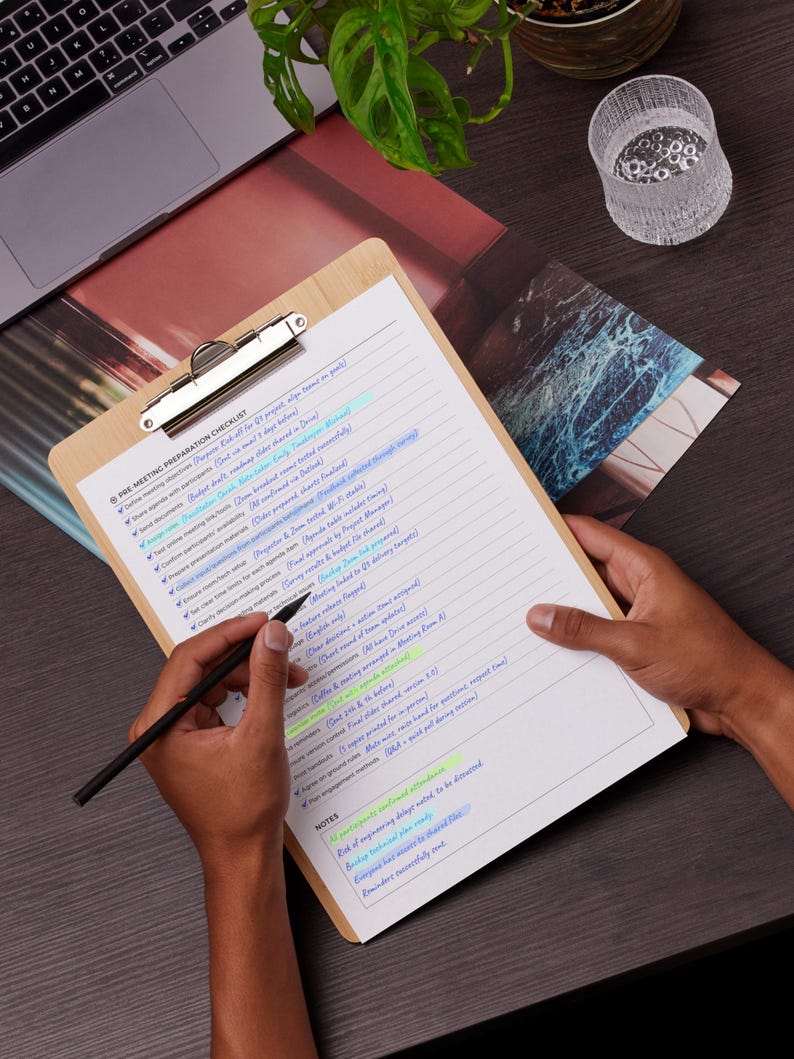 May include: A wooden clipboard with a checklist titled "PRE MEETING PREPARATION CHECKLIST" is held by a person. A black pen is poised over the paper. The checklist has highlighted text in blue and yellow. A laptop and a glass are in the background.