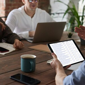 May include: A meeting is taking place around a wooden table. A tablet displaying a document is held by a person, while others use a laptop and a smartphone. A teal mug of coffee and a black smartphone are on the table.