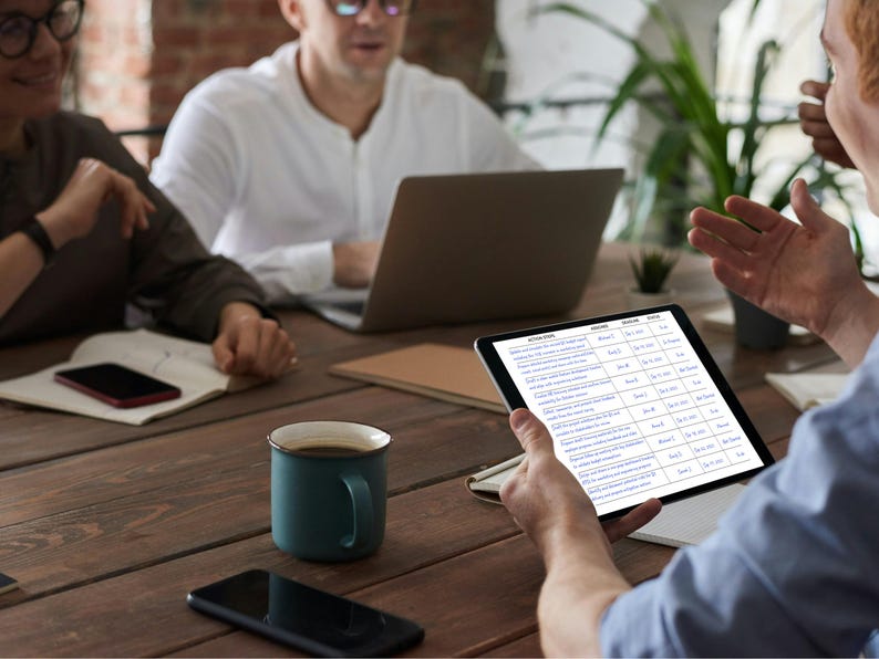 May include: A meeting is taking place around a wooden table. A tablet displaying a document is held by a person, while others use a laptop and a smartphone. A teal mug of coffee and a black smartphone are on the table.
