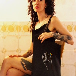 A funky woman with tattoos sits in a setting with a cool vibe. She wears a black tote on her shoulder, on it, a rainbow heart is pinched between two fingers of a white, line drawn hand