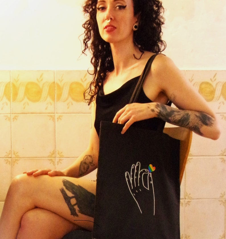 A funky woman with tattoos sits in a setting with a cool vibe. She wears a black tote on her shoulder, on it, a rainbow heart is pinched between two fingers of a white, line drawn hand